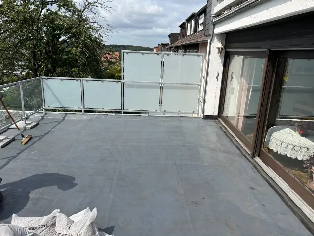 Große Dachterrasse mit Glasgeländer im Vorzustand: graue Abdichtung/Untergrund, Besen und Materialsäcke am Rand.