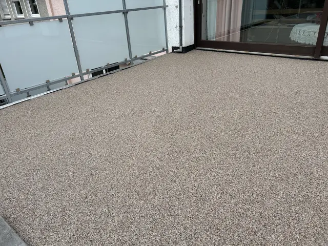 Fertig beschichtete Terrasse mit beigem Steinteppich, fugenlos bis an Türschwelle und Geländeranschlüsse verlegt.