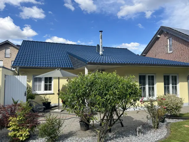 Gelbes Einfamilienhaus mit frisch gereinigtem, blau beschichtetem Ziegeldach und gepflegtem Garten unter blauem Himmel.